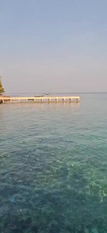 A video captures a serene ocean scene with a clear, shallow water revealing the sandy bottom. A white pier extends into the turquoise water on the left side of the frame. In the distance, a small boat can be seen on the horizon. The sky is a pale blue, suggesting a calm day. The water has a gentle ripple effect, and the sunlight filters through it, creating shimmering patterns on the seabed. The overall atmosphere is peaceful and tranquil.