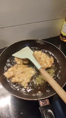A light green spatula is used to flip golden brown fried food in a black frying pan. The food appears to be cut into irregular pieces and is submerged in bubbling oil. The pan is on a black stovetop. The background is a gray backsplash and a partial view of a bottle of oil with a yellow label. The camera is positioned above and slightly to the side of the pan.