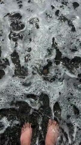 The video shows a pair of bare feet standing on a dark, pebbly shore. The waves are gently washing over the pebbles, creating white foam. The water is clear and shallow. The feet are positioned in the center of the frame, with the toes pointing forward. The pebbles are of various sizes and colors, with some larger white and tan rocks mixed in with the darker ones. The overall impression is one of a serene beach scene.