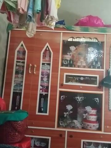 The video shows a cluttered room with a wooden wardrobe as the focal point. The wardrobe has glass doors with decorative patterns and drawers. On top of the wardrobe are stacked colorful fabrics and what appears to be a pink balloon. To the left of the wardrobe, a stack of folded clothes is visible. The room's walls are painted gray, and a patterned ventilation opening lets in light. The camera is positioned at a low angle, looking up at the wardrobe and the items around it. The scene suggests a domestic setting, possibly a bedroom or storage area.