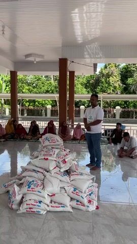 The video shows a community gathering in an open, covered area with a reflective tiled floor, likely a veranda or the entrance to a mosque. A large group of predominantly elderly women, dressed in colorful hijabs, are seated on the floor in neat rows, facing forward. In the foreground, a substantial pile of white sacks, labeled "Beras Tanggamus Pulen" (Tanggamus Pulen Rice), is visible. Among the seated women, a cardboard box with "Dua air" printed on it can be seen. On the right side of the frame, a man stands, wearing a white polo shirt and jeans, with a white tag or badge on his chest displaying "DNI." The background features a white building with arched windows, typical of mosque architecture, and lush green trees under bright daylight, indicating a warm, sunny climate. The scene suggests a food aid or charity distribution event, with the women attentively waiting for or listening to instructions.