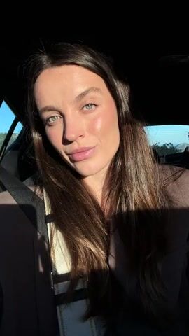 The video features a woman sitting in a car. She has long, dark hair, light eyes, and is wearing a brown blazer. She is wearing a seatbelt. She is looking at the camera and smiling. The light changes in the video, sometimes illuminating her face and sometimes casting it in shadow. Through the car window, we can see a glimpse of a green landscape. Music is playing in the background. She shows a ring.