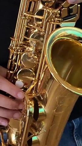 A close-up shot shows a pair of hands playing a shiny, gold saxophone. The fingers are shown pressing down on the keys of the instrument, producing musical notes. The background is dark, with the exception of some green accents on the saxophone itself. The video appears to be a short, isolated segment of a musical performance.