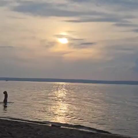 A video shows a person partially submerged in water near a sandy shore, with a body of water stretching to the horizon. The sun is visible in the sky, partly obscured by clouds, casting a reflection on the water's surface. The scene appears to be at dusk or dawn, with muted lighting.
