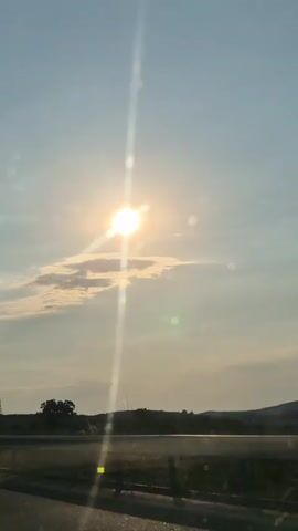 This video captures a daytime view from a moving vehicle on a highway. The sun is high in a clear blue sky, casting a bright, lens-flared glow that dominates the frame. Wispy white clouds drift across the sky, partially obscuring the sun at times. In the distance, a flat landscape is visible, with a dark line of trees and rolling hills forming the horizon. A tall, latticed power pylon stands prominently on the left side of the frame, with power lines stretching across the scene. The bottom of the frame shows a glimpse of the road and roadside vegetation, indicating the perspective of someone traveling. The overall impression is one of a bright, sunny day, likely late morning or early afternoon, suggesting travel through a rural or semi-rural area. There is no visible text in the media file. The serene natural scenery and the bright sunlight evoke a sense of openness and peaceful travel.
