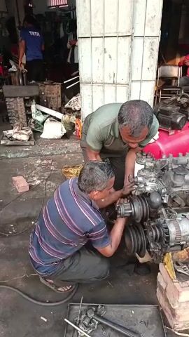 The video shows the interior of an automobile repair shop. Two men work on an engine block in the foreground. Another man inspects a second engine block further back in the shop, using a flashlight to illuminate the interior. The shop is cluttered with tools, machinery, and multiple engine blocks in various states of repair. The floor is stained and oily. The entrance to the shop is a large metal grid door. The overall impression is of a busy, functional workspace dedicated to engine repair.