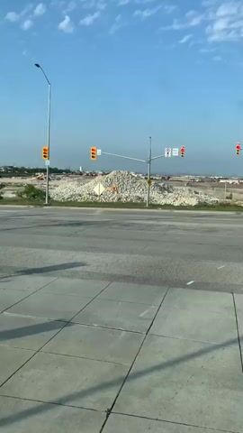 The video shows a construction site with a large pile of rubble in the background. There are traffic lights and signs visible. A large red and black semi-truck is in the foreground, driving across the intersection. The sky is blue with some clouds. The ground is paved with asphalt.
