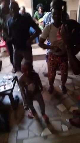 The video shows an indoor scene with several people of varying ages dancing to music. There is a small child in a grey outfit, facing the camera while people dance around him. A woman in a grey shirt and patterned pants is standing with her hands on her hips. A man in a red shirt and blue shorts is crouching. The flooring is tile. There are various objects scattered around the room, including chairs, shoes, and debris. The lighting is somewhat dim, and the camera seems to be handheld, resulting in some shakiness.