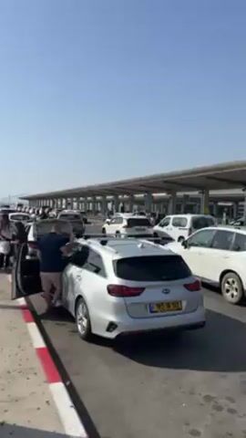 The video captures a busy scene at what appears to be an airport drop-off area on a sunny day.  Several white and silver vehicles are lined up, many with Israeli license plates. People are getting out of cars, some retrieving luggage. One man is pulling a suitcase across the crosswalk towards the covered terminal area, where columns support a roof with many lights and cameras. The road is marked with a red and white stripe along the edge.