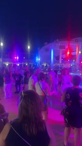 A group of people are dancing on a wooden deck at night. The scene is lit with purple and blue lights. Some people are wearing casual clothing, while others are dressed up. The people are of various ages and ethnicities. Some are dancing alone, while others are dancing in pairs or groups. There are some tables and chairs in the background. The atmosphere appears to be festive and lively.