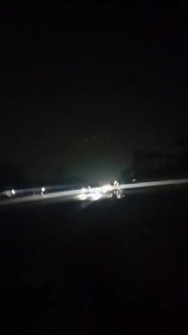 The video shows a nighttime view of a road with vehicles passing by, indicated by streaks of light from their headlights and taillights. The environment appears to be dark with some trees or foliage visible on the sides of the road. The ground seems wet, possibly from rain, reflecting the vehicle lights. The camera is in motion, creating a sense of movement and dynamism in the scene.