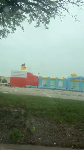 The video shows a view of a large inflatable attraction, possibly a bouncy castle or inflatable obstacle course, situated in an outdoor area. The inflatable structure is predominantly white, red, and light blue, featuring cartoonish duck figures with hats. To the left of the inflatable, a portion of a red slide or ramp is visible. The inflatable appears to be in a paved area, possibly a parking lot, with some debris scattered on the ground. In the foreground, a tree with green leaves is partially obscuring the view, and the surface in front of the tree appears to be dirt and grass. The sky is overcast and appears to be raining, as evidenced by raindrops visible on the camera lens or windshield. The overall impression is of an outdoor amusement setup experiencing inclement weather.