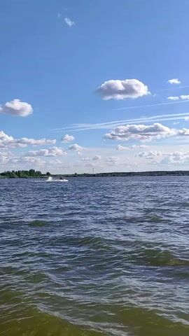 The video shows a person riding a jet ski on a body of water. The jet ski is creating a spray of water as it moves. The sky is blue with some white clouds, and there is land in the background with some trees and a tower. The water is choppy.