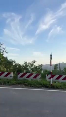 The video depicts a roadside view from a moving vehicle. The road has a grey surface with a white painted line separating lanes. Alongside the road, there is a concrete barrier painted with alternating red and white diagonal stripes. Beyond the barrier, there is sparse green vegetation. In the distance, a hazy, mountainous landscape is visible under a bright, clear blue sky with wispy cirrus clouds. Tall grasses sway in the breeze.