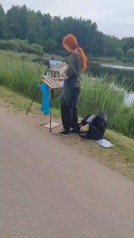 A person with red hair, wearing a dark apron and dark pants, stands by a lake. They are painting on an easel. Beside them is a backpack and a palette of paints. The scene is outdoors with trees and water in the background.