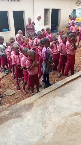 A group of young children, all in school uniforms, are standing in a line outside a building. The children are mostly wearing red and pink uniforms, with a few wearing other outfits. The children are standing with their heads bowed or their hands covering their faces. There are two adults standing in the background, and a yellow school bus is visible in the distance. A woman in a black shirt and red skirt is standing to the side of the children, with her back to the camera. The children are all very young. The environment appears to be a schoolyard. They are praying.