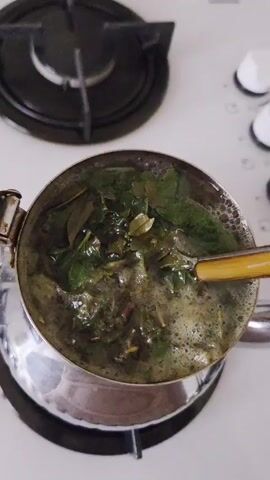 The video shows a silver Turkish teapot on a white gas stove. Inside, green leaves and water are being stirred with a small wooden spoon, producing steam and bubbles. The stove has a black burner next to the teapot.