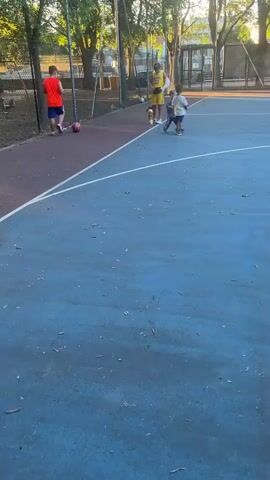 A group of four people, including three children and one adult, are on a blue and red outdoor court. The adult, wearing a yellow shirt and shorts, is bending over a golden trophy that sits on the ground. Two young children are standing near the trophy, one in a white shirt and the other in a blue shirt. A third child, wearing a red shirt and blue shorts, stands further away on the red part of the court, next to a pink soccer ball. The scene appears to be a park or recreational area, with trees and a fence visible in the background. The lighting suggests it is late afternoon or early evening, with the sun casting long shadows. The overall atmosphere seems relaxed and playful, with the children engaged in an activity around the trophy.