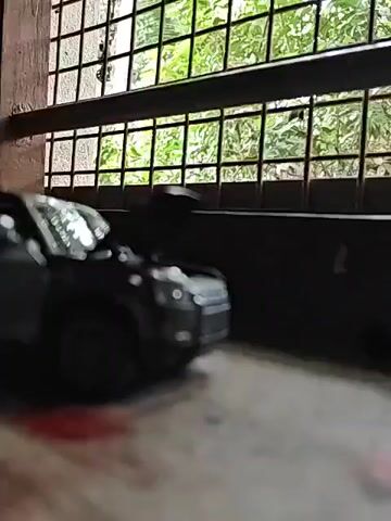 A dark-colored toy SUV is positioned on a surface, possibly a windowsill, with a window behind it showing greenery and a grid-like structure. The SUV's hood and driver's side door are open. Red liquid, possibly paint or dye, is visible on the surface in front of the SUV. The camera angle is low, focusing on the toy vehicle.