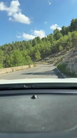 The video shows a view from the dashboard of a car driving on a winding road through a wooded, hilly area. The road is paved and has white lane markings. The car is moving forward, and the camera is pointed out the windshield. The sun is shining, and there are some clouds in the blue sky. The hillsides are covered in green trees and shrubs, with some rocky outcrops. The video is from the perspective of the driver.