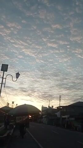 The media file depicts a street scene at sunrise in Desa Toto Selatan, Indonesia. The sky is filled with a dense pattern of altocumulus clouds, illuminated by the soft glow of the rising sun peeking through the clouds on the left side of the frame. The light creates a warm, golden hue in the sky, contrasting with the pale blue above.

The street itself appears to be a two-lane road, with buildings lining one side. These structures seem to be a mix of homes and small businesses, with some featuring covered stalls or awnings. A few utility poles and power lines are visible, indicating a populated area. In the foreground, a motorcycle is parked near a stall with a green umbrella, suggesting it might be a vendor or a customer. Further down the street, a black pickup truck is driving away from the camera, its taillights reflecting in the dim light. A vintage-style street lamp with a curved arm stands on the left side of the road.

The overall mood is peaceful and serene, capturing the quiet start to the day. The presence of the clouds suggests it may be humid, typical of tropical weather. The scene is a glimpse into the daily life of a village in Indonesia as it awakens. There is no discernible text visible in the media file.