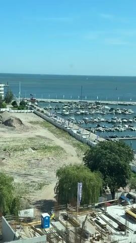 The video pans across a marina on a sunny day, showing numerous boats docked in the water. In the foreground, there's a construction site with scaffolding and building materials, including a portable toilet. Beyond the construction, there are trees and a paved area with people walking. The marina itself is filled with various types of boats, from small speedboats to larger yachts and sailboats. The water is calm, and the sky is clear blue with a few distant boats visible on the horizon.