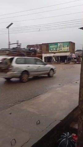 The video shows a street scene with several vehicles and buildings. In the foreground, a wet, paved road is visible, with a metal manhole cover. In the background, there are several buildings, some appearing to be shops or residences. A street lamp is visible. The sky is overcast.

Several vehicles are seen moving on the road. At the beginning of the video, a silver station wagon is visible, with its trunk open and loaded with items. A black SUV then passes by. Following that, a motorcycle with two occupants and a large, flat object strapped to the back travels down the road. A white car is parked on the side of the road in the distance. Various objects, including tarps and debris, are scattered along the side of the road. The overall impression is a busy street in a somewhat unkempt urban environment.