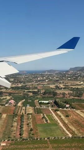 The video shows a view from an airplane window during a landing. The plane's wing, with a blue tip, is visible in the foreground. Below, the landscape consists of fields, trees, and scattered buildings. In the distance, there are mountains and the sea. The sky is clear and blue. The general impression is of a sunny day over a rural or semi-rural area, likely near the coast.