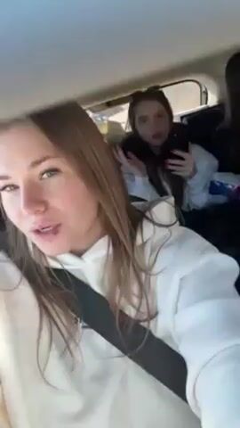 Three young women are inside a car. The woman in the front seat is filming with a handheld camera, and the two other women are seated in the back. The front passenger is wearing a white hooded sweatshirt and a seat belt. The two women in the back are sitting up, but it's hard to see them clearly. Music is playing in the car. The car seems to be moving.