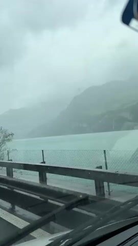 The video captures a view from inside a moving vehicle, likely a car or bus, on a rainy, overcast day. Through the wet windshield, with wipers actively clearing the rain, a mountainous landscape with a large, turquoise body of water is visible. The vehicle appears to be traveling on a road or highway next to the water, with a guardrail and chain-link fencing separating the road from the lake. Lush green trees, some swaying slightly, line the road, while towering mountains, partially obscured by mist and heavy clouds, rise in the background. The sky is uniformly grey. The overall atmosphere is damp and misty, characteristic of a rainy day in a mountainous region like Glarus Nord, Switzerland. A small, indistinct object, possibly an air freshener or ornament with an unreadable logo, hangs from the rearview mirror area. No people are visible.