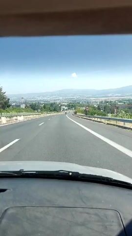 The video presents a first-person view from inside a vehicle moving forward on a paved two-lane road under a clear blue sky on a sunny day. The dark dashboard and lower edge of the windshield are visible at the bottom of the frame. White lane markings delineate the road. A white guardrail lines the right side of the road, bordered by green vegetation including trees and shrubs. In the mid-distance on the right, a circular red-bordered speed limit sign displays "90". Further in the distance, an expanse of buildings indicates an urban or suburban area, backed by a range of mountains. The view consistently moves forward throughout the video.