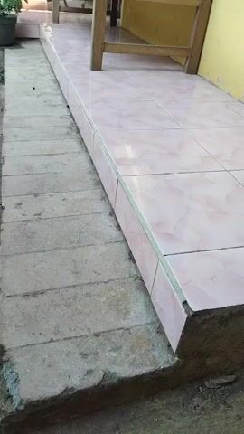 The video shows a tiled walkway leading to a house. A wooden bench sits on the walkway. There are plants in pots on either side of the walkway. A motorcycle is parked in front of the house. The house is painted yellow. The video pans from left to right, showing the walkway and the house in more detail.