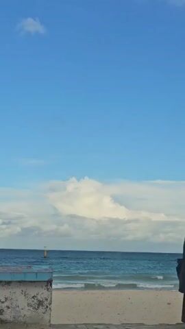 A scenic view of a beach with a person standing on the right side of the frame. The beach has light-colored sand, and the ocean extends to the horizon, where it meets the blue sky. White, fluffy clouds dot the sky, creating a picturesque backdrop. A small, yellow buoy is visible in the water near the horizon. In the foreground, on the left, there is a low, concrete structure with a weathered, textured surface. The person on the right is dressed in dark clothing.
