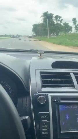The video shows a first-person perspective from inside a car, looking through the windshield at a road with a group of people and several vehicles. The scene appears to be a roadside in Kwali, Nigeria. The weather is overcast, suggesting a cloudy or possibly rainy day.

In the distance, there are multiple cars and what look like tires or barricades on the road, indicating some sort of disruption or gathering. Closer to the car, there are several people standing around. Some appear to be talking or observing the situation. The vehicles visible include cars, vans, and possibly trucks. One van in the middle distance has what looks like a red light on its roof, perhaps indicating an official vehicle.

The interior of the car is partially visible, including the steering wheel and the car's dashboard with its controls and display screen. The screen shows "Bluetooth Audio" and a music playback interface, suggesting the driver is listening to music or a podcast. The overall impression is one of a slow-moving or stopped traffic situation, with people gathered on the side of the road, possibly due to a checkpoint, a roadblock, or a social event. There are no overt signs of distress or panic, but the presence of the barricades and the gathering of people suggests an unusual event.