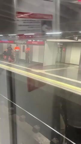 This video shows a subway platform with several people. In the foreground, there is a reflection of a glass window with railing. On the platform, there are four individuals wearing uniforms. Two of them are wearing orange vests and appear to be law enforcement or emergency personnel. One is wearing a dark uniform with "POLICE" visible on the back. Another person, dressed in civilian clothing, stands near a bench. There is a stretcher or medical equipment on the ground with several people gathered around it, appearing to attend to someone. The platform has striped markings on the floor and signs indicating a station name "Terrassa." A fire extinguisher sign is visible on the wall. A poster with text is hanging above the platform.