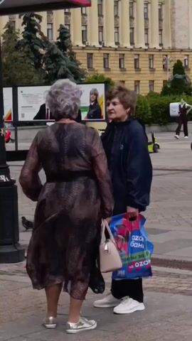 The video shows two elderly women standing on a paved outdoor area, possibly a plaza or sidewalk. The woman on the left has short, gray, curly hair and is wearing a long, dark, patterned dress and silver sneakers. She is holding a light-colored handbag in her left hand. The woman on the right has short, brown hair and is wearing a dark blue jacket, black pants, and white sneakers. She is holding a blue shopping bag with red handles in her right hand. The women are facing each other and appear to be engaged in conversation. In the background, there is a building with multiple windows, a large billboard with images of people, and other individuals walking in the distance. The overall environment appears to be urban.