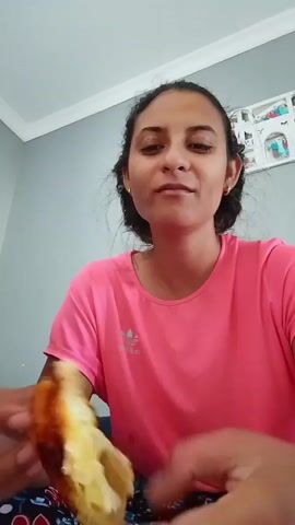 A young woman with dark hair and a pink t-shirt is sitting in front of a gray wall, eating a pastry. She is holding the pastry in her hands and taking a bite. She appears to be enjoying the pastry. The room is sparsely decorated with a framed picture on the wall.