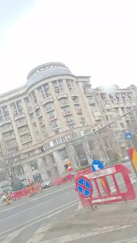 The video shows a view from inside a car driving on a road beside a large, multi-story building. The building is made of light-colored stone or concrete and has many windows and balconies. It also has a rounded structure on top. There are trees along the roadside, and some road construction is underway with orange barriers. Several cars are parked along the side of the road and moving in the road. The sky is overcast and gray.