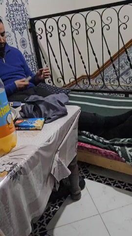 A man with a beard and a blue sweater sits on a bench in front of a staircase with black railings. He is holding a cigarette and a lighter, preparing to light the cigarette. A small table next to the bench holds a bottle of juice, a box of cookies, and a gray garment. The floor is tiled in a black and white pattern, and the wall behind the staircase has blue and white decorative tiles.