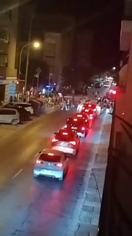 The video shows a nighttime street scene with several cars in a line, their brake lights illuminated. A group of people are visible on the sidewalk to the left, some appearing to be watching the cars. In the background, there are buildings and streetlights. The perspective is from a higher vantage point, looking down onto the street. There appears to be a police car with blue lights visible further up the street. One person on the sidewalk is holding up a phone, possibly recording the event.