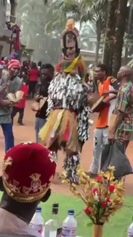 The video captures a scene of a cultural celebration. A person in an elaborate masquerade costume, adorned with silver strips and yellow fibers, is the central figure. They are surrounded by a group of people, some of whom are dancing and playing musical instruments, including a drum. One person is seen holding and handing out woven hats. The foreground features the back of someone wearing a red velvet hat with gold trim. A table with decorative items, including a vase of artificial flowers and bottles, is also visible. The setting appears to be outdoors, with trees and buildings in the background. The environment suggests a cultural or religious event with traditional music and attire.