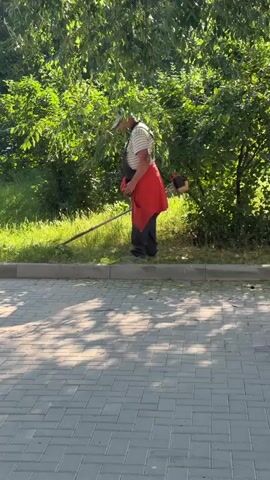 A person wearing overalls and a red jacket over a striped shirt is mowing grass with a string trimmer. The person is standing on a paved area next to a strip of grass. Trees and bushes are visible behind the person. The sun is shining, casting shadows on the ground.
