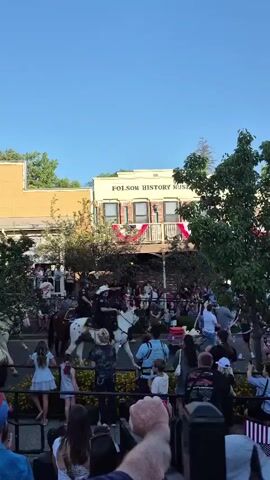 A video shows a parade with people watching from the sidewalk in front of the Folsom History Museum. Several horses with riders are visible in the street, and a large tree stands in the foreground. The sky is clear and blue, suggesting a daytime event. People are dressed in casual clothing, and some are holding American flags. The overall atmosphere appears festive and celebratory.
