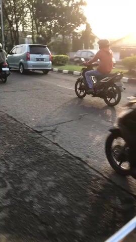 This video captures a street scene in Candi, East Java, during what appears to be late afternoon or early evening, given the warm, soft light and long shadows. The weather seems pleasant, likely dry, as the road surface is visible and there's no indication of rain.

The primary subjects are several individuals riding motorcycles. In the foreground, the rear view of a person in a dark jacket and helmet is shown, riding a dark-colored motorcycle. Further down the road, another motorcycle is visible with a rider wearing a colorful, patterned shirt and a red helmet, carrying large white bags on the back. The motorcycles are traveling on an asphalt road with a distinct crack running across it. The road is bordered by a sidewalk with alternating black and white markers.

Beyond the sidewalk, there is lush greenery, including trees and manicured bushes. In the background, a few white vans are parked or driving, suggesting a public area or commercial zone. A building with a white facade is partially visible behind the trees. There's also a glimpse of what might be a person walking or sitting on a bench in the distance. The overall atmosphere is tranquil and everyday, suggesting a normal commute or delivery activity. The golden light filtering through the trees creates a slightly nostalgic or peaceful mood.

There is some visible text on the license plate of the second motorcycle, which appears to be "JX 2285".