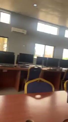 The video shows an interior view of a computer lab. There are multiple rows of desks, each equipped with a computer monitor, keyboard, and mouse. The desks are made of brown wood. Blue chairs with a gold trim are placed in front of each desk. The walls are painted a light gray color. Several windows with metal bars cover the outside views. The room is well lit by ceiling lights and natural light coming through the windows. An air conditioner is mounted on the wall. The video is taken from a static position, looking over a desk. There are no people visible in the video.