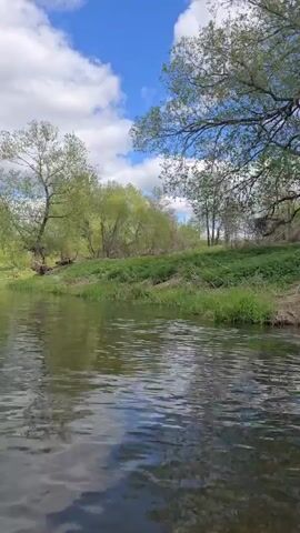 The video shows a person kayaking on a river. The camera is mounted on the front of the blue kayak, looking forward. There are green trees on either side of the riverbank. The sky is blue with scattered white clouds.  In the distance, another person is kayaking in a red kayak. The water surface is calm with ripples.