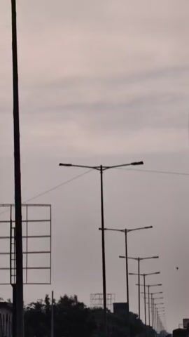 The video shows a view from a moving vehicle on a highway. A line of tall lampposts with multiple lights stretches along the right side of the road. On the left, there's a yellow fence, trees, and a large billboard structure. The sky is overcast. Several vehicles, including cars and trucks, are visible on the highway, moving away from the camera. The road has clear white lane markings.