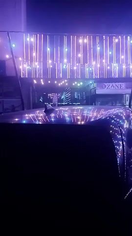 Okay, here's a detailed description of the video:

The video is shot at night and focuses on a dark-colored car parked on a street. The reflection of the street lights is visible on the car's roof and side, creating a sparkling effect. Across the street, a building is adorned with bright, vertically-oriented lights. These lights are predominantly white with touches of blue.

The name "OZONE" is visible on the building's facade. Further down the street, there appears to be a Christmas tree or similar decorative tree illuminated with lights. A figure can be seen walking in the background.

The overall atmosphere is dark with a strong emphasis on the illumination provided by the streetlights and building decorations. The focus is on the visual contrast between the darkness and the bright lights reflecting off the car.