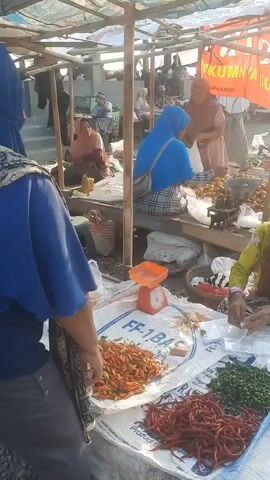 A busy outdoor market scene. In the foreground, a person wearing a blue headscarf and a patterned vest faces away from the camera. In front of them, on a white tarp, are piles of red chilies and green peppers, along with a red and orange scale. To the right, another person is partially visible, appearing to be handling some items. In the background, several stalls are set up, with people browsing and interacting. One stall has a large orange banner with text that is partially obscured. The market appears to be under a makeshift roof supported by poles. The overall environment suggests a lively marketplace, possibly in a rural or semi-urban setting.