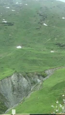 The video shows a lush green mountainside in a cloudy or misty setting. The camera pans slightly to show the landscape, including some patches of snow near the top of the mountain. A road and railing are visible at the bottom of the frame, and a road sign indicating a curve appears briefly. The overall scene is natural and tranquil.